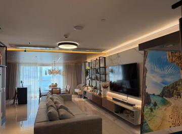 Living do apartamento de 196 m² no One Tower, com painel de TV e o mar ao fundo · Apartamento decorado com 4 suítes e vista mar no One Tower, Barra Sul